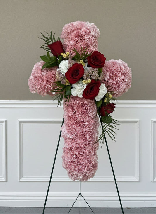 Floral arrangement in the shape of a cross with pink and red flowers on a stand against a beige wall.
